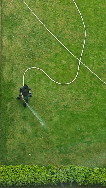 An aerial view of a gardener in a dark uniform, watering a well-maintained, lush green lawn with a garden hose connected to a long white hose reel. The lawn features a smooth, even surface with visibl