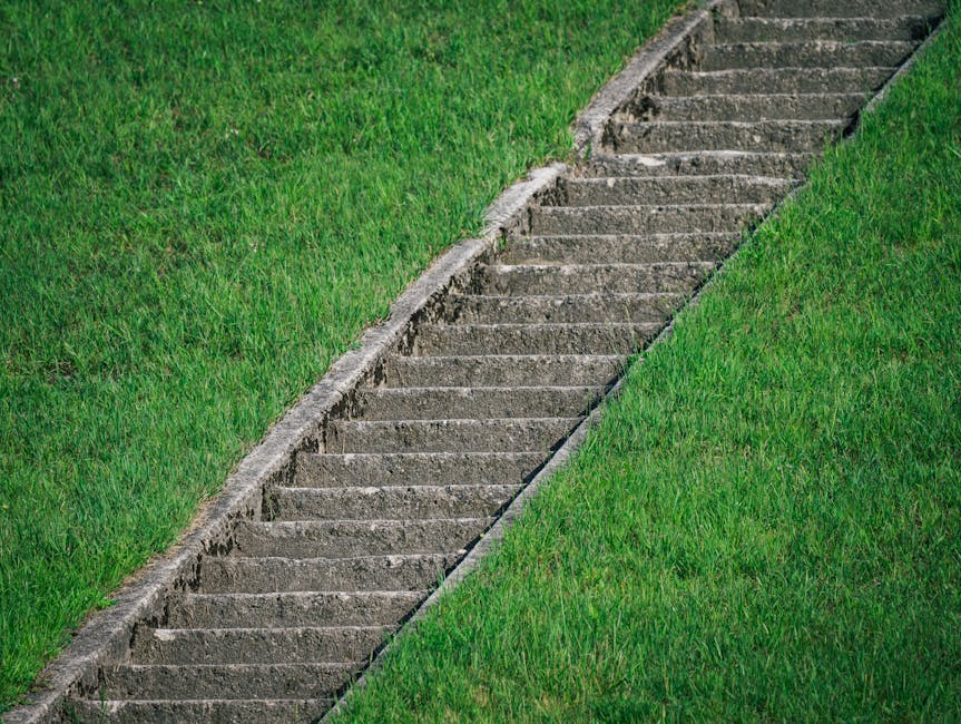 An aerial view of a gardener in a dark uniform, watering a well-maintained, lush green lawn with a garden hose connected to a long white hose reel. The lawn features a smooth, even surface with visibl